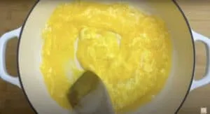 Stirring eggs in a white pot with wooden spatula, creating a swirl pattern for scrambled eggs.